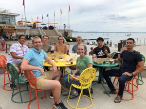 Group of students with their professor sitting in colorful chairs outside at a food event. 