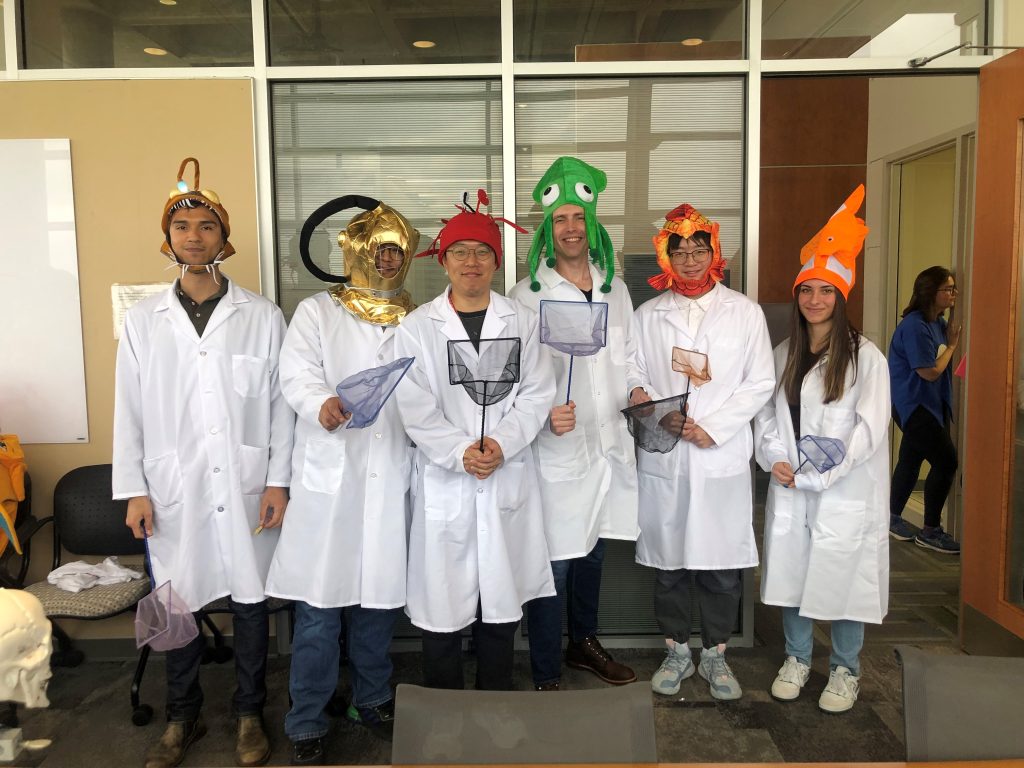 Four students in lab coats wearing colorful fish hats for a Halloween party. 